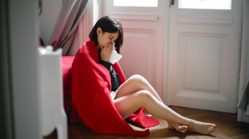 A person wrapped in a blanket sitting on the floor, blowing their nose.