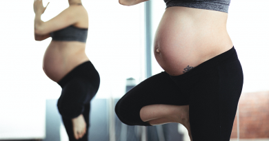 A pregnant person with a large, exposed stomach stands in a yoga pose - hands clasped at shoulder height, one leg tucked into the other.